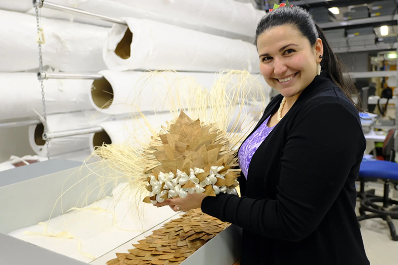 A woman is holding a headdress made of pandanus leaves and shells and smiling at the camera.