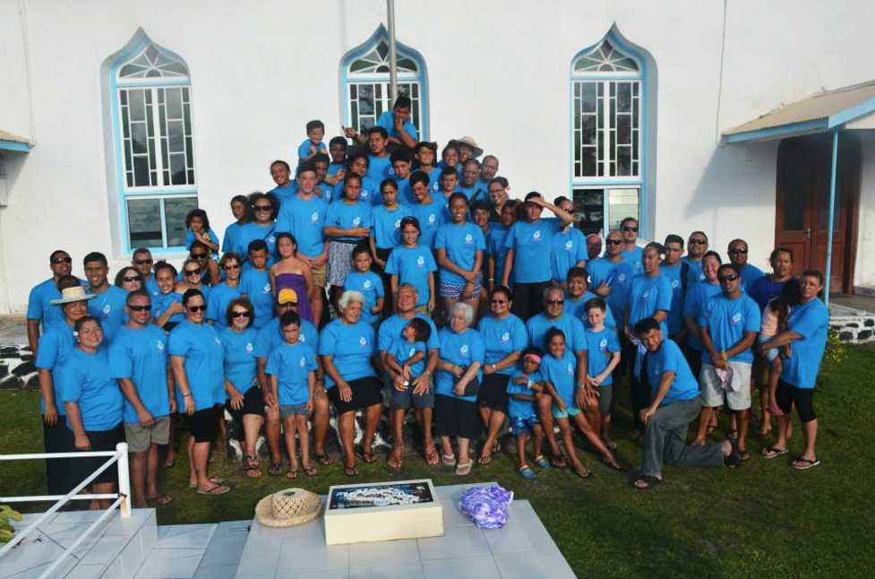 A large group of people are assembled in front of a church. Nearly all of them are wearing the same bright blue coloured shirt.