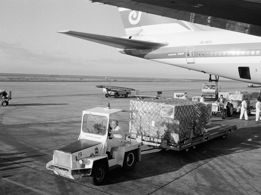 Te Māori exhibition, Brian Brake; photographer; about 1986; New Zealand A black and white photo of an airport runway with part of a plane and a luggage transport cart and trailer in the foreground
