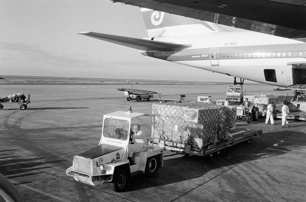 A black and white photo of an airport runway with part of a plane and a luggage transport cart and trailer in the foreground