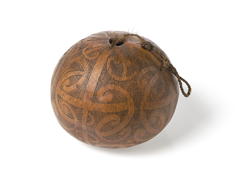 A roung gourd that has carved Māori designs all over it and a cord attached via holes at the top.