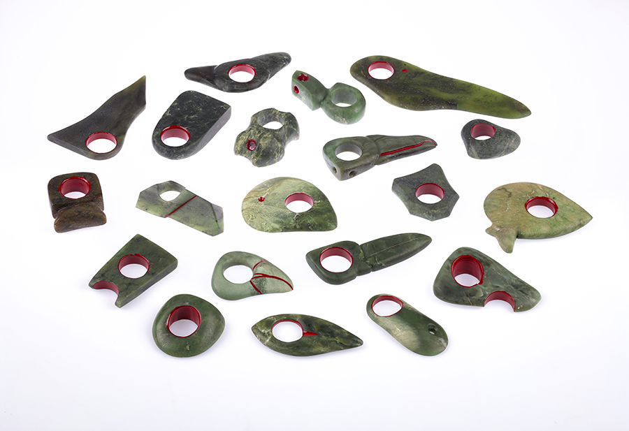 Many pieces of greenstone with a single hole in each that turns them into rings with a red marking are laid out on a white surface.