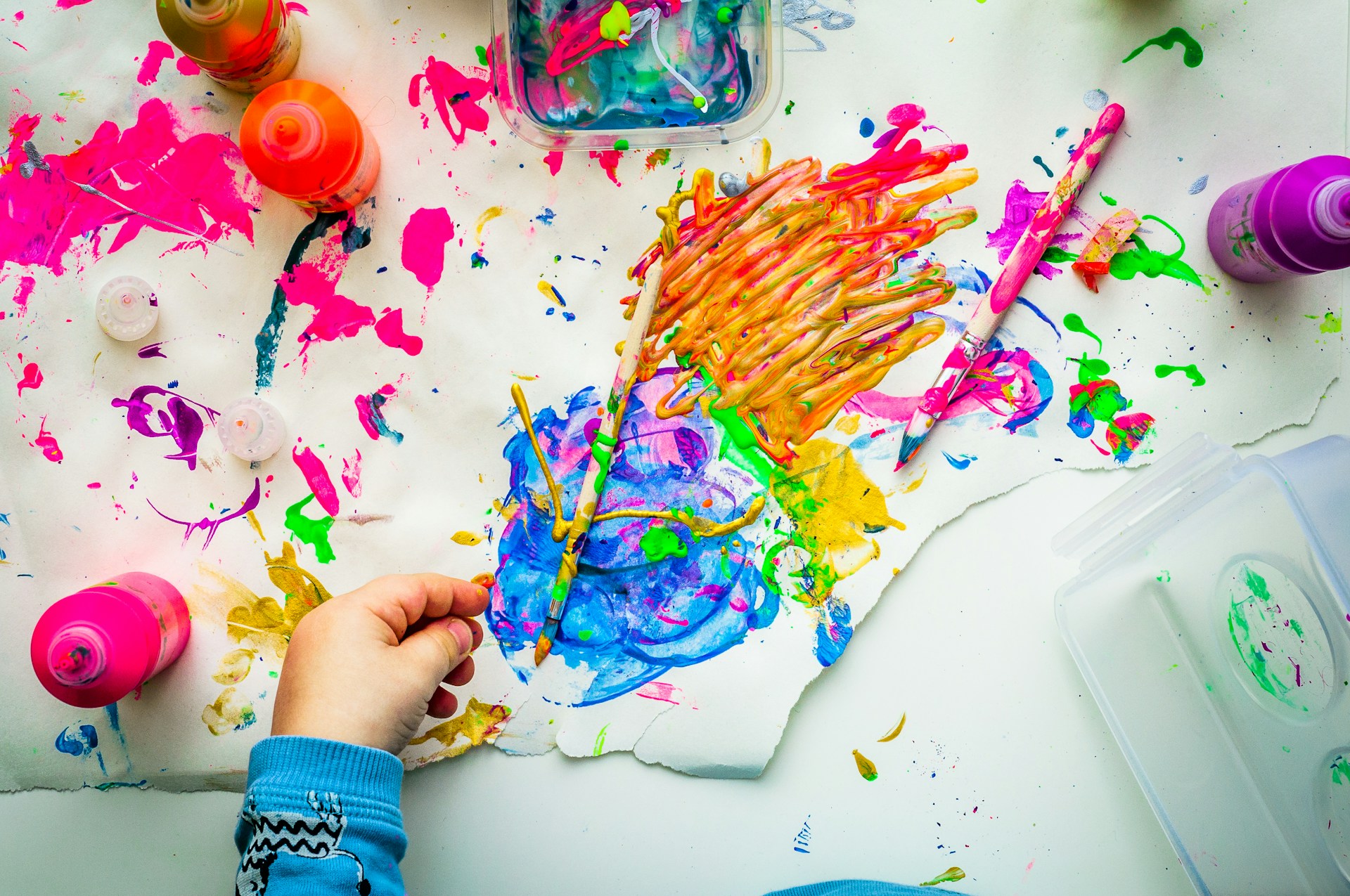 A photograph of a desk with a lot of paint colours everyone and one hand resting on the desk.