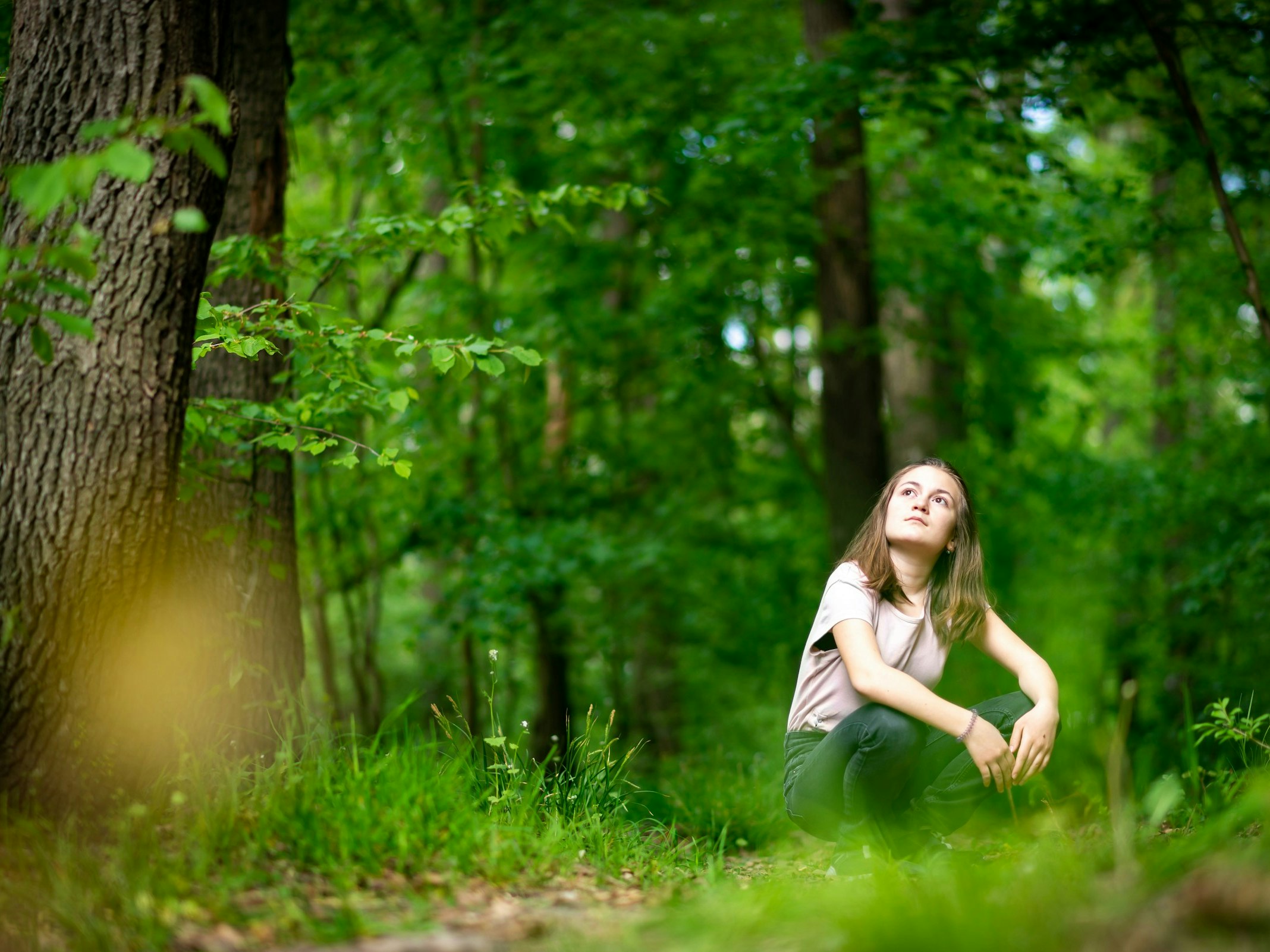 Photo by Michal Vrba. Unsplash A young woman is sitting on the ground in a forest. She is looking up at the trees.