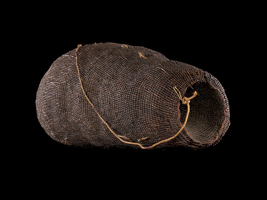 Hinaki / Eelpot, date and maker unknown. Te Papa (ME022717) A woven basket with a plaited handle sitting in a black background.