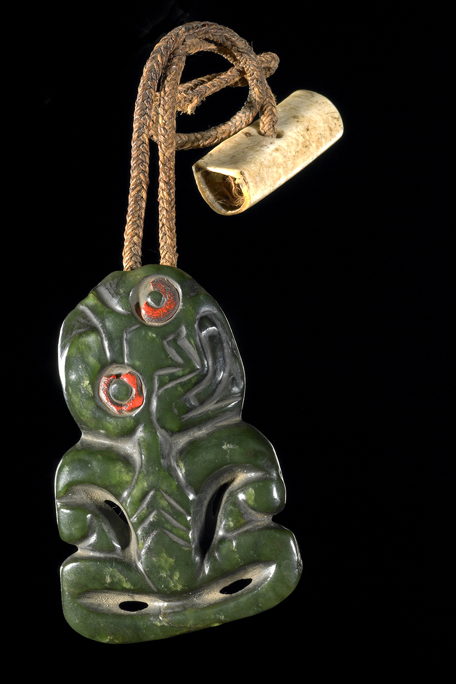 A pounamu tiki or greenstone pendant in a stylised human form with red tinted eyes. It is hanging from a cord and is sitting on a dark background.