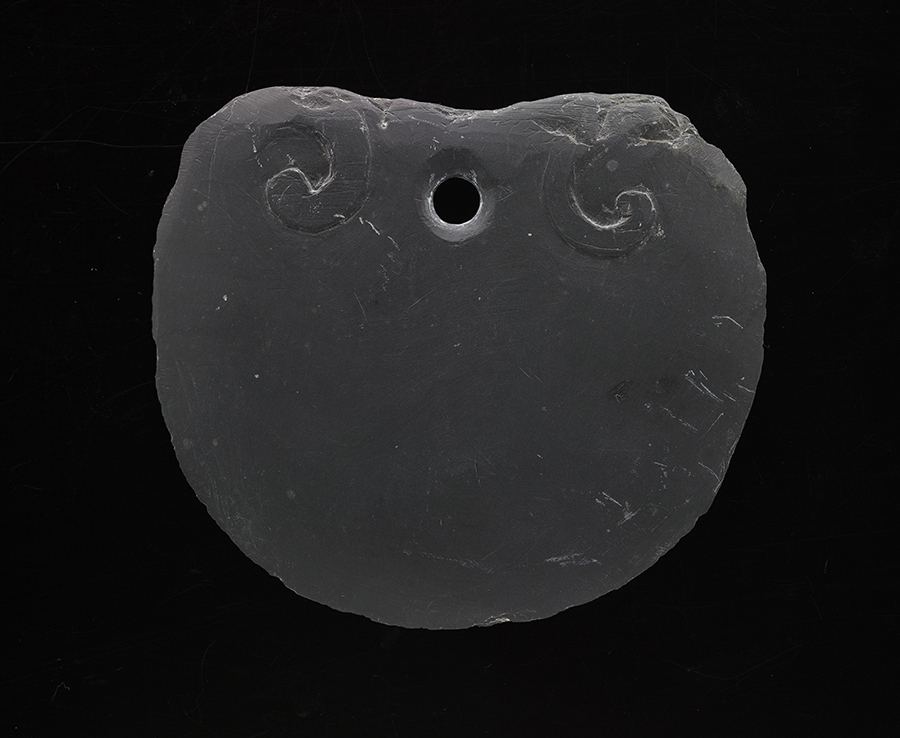 A piece of flat slate with small carved designs at the top along with a hole for a cord.