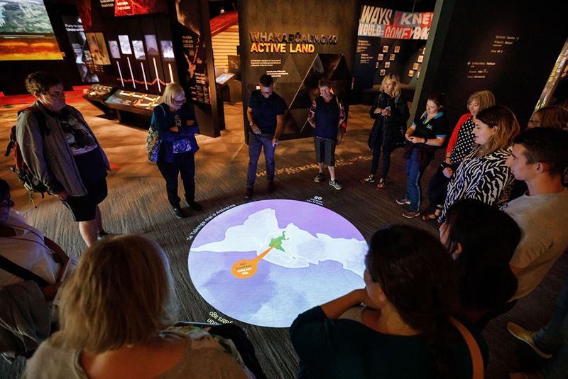 A lot of people standing around a floor projection of part of the world map on the floor.