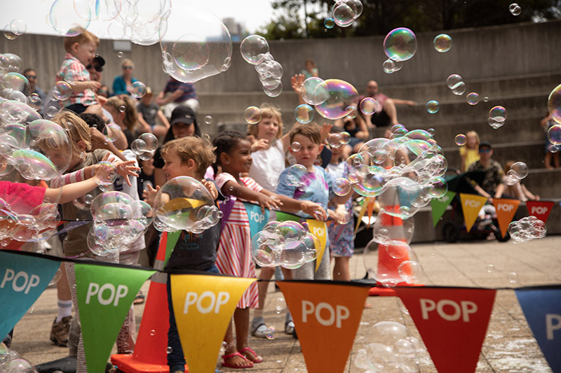 Lots of children are playing together outside. There is a banner with the word Pop on each of the small flags and there are bubbles everywhere.