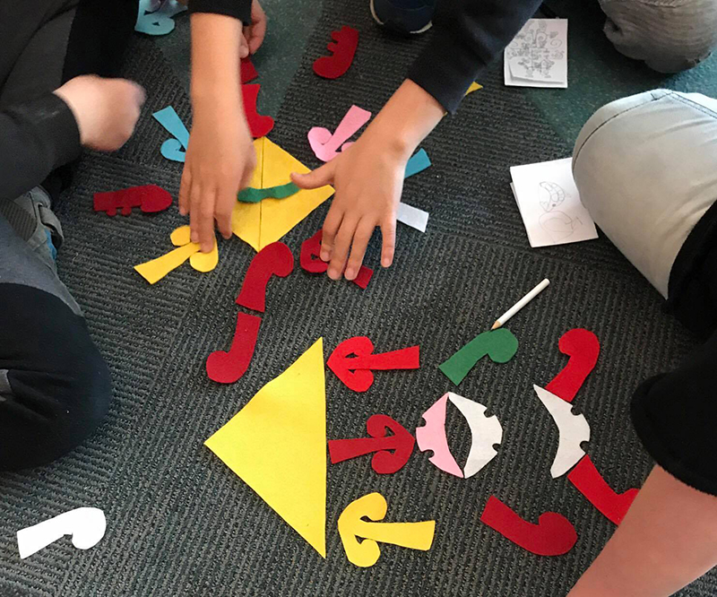 A view of cut out paper shapes on a floor with a knee and several hands in shot.