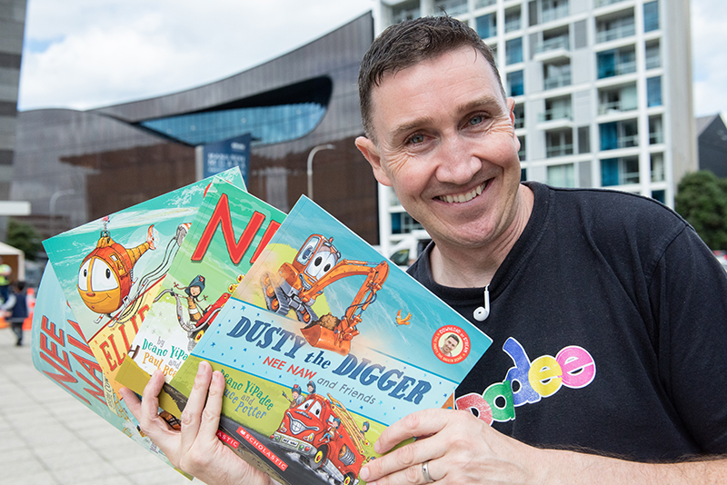 Asmiling man is standing outside and holding up four flat children's picture books.