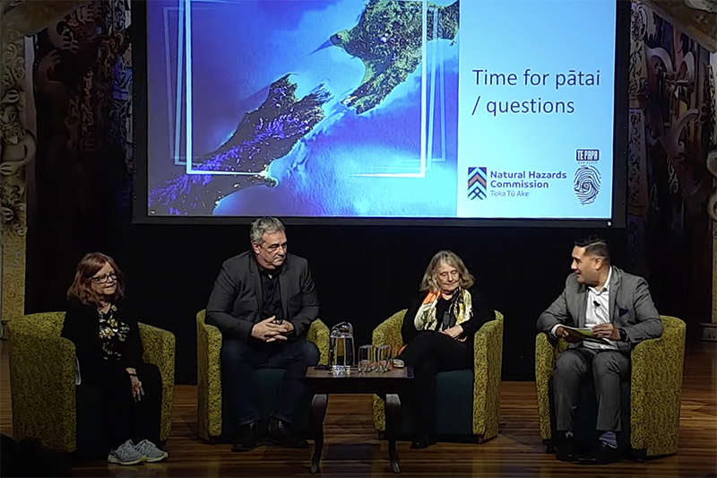 Two men and two women are sitting on a stage talking to an unseen audience. There is a large projection screen behind them.