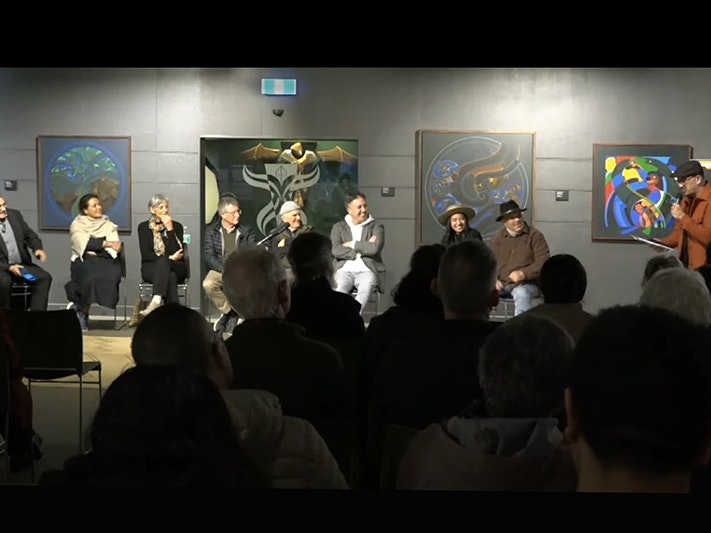Still from Turanga FM video of Te Maori panel talk. Eight people are sitting in front of an audience while one man is standing at the end talking to them. They are all smiling.