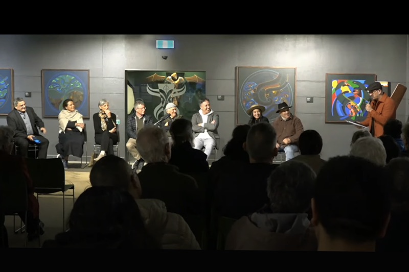 Eight people are sitting in front of an audience while one man is standing at the end talking to them. They are all smiling.