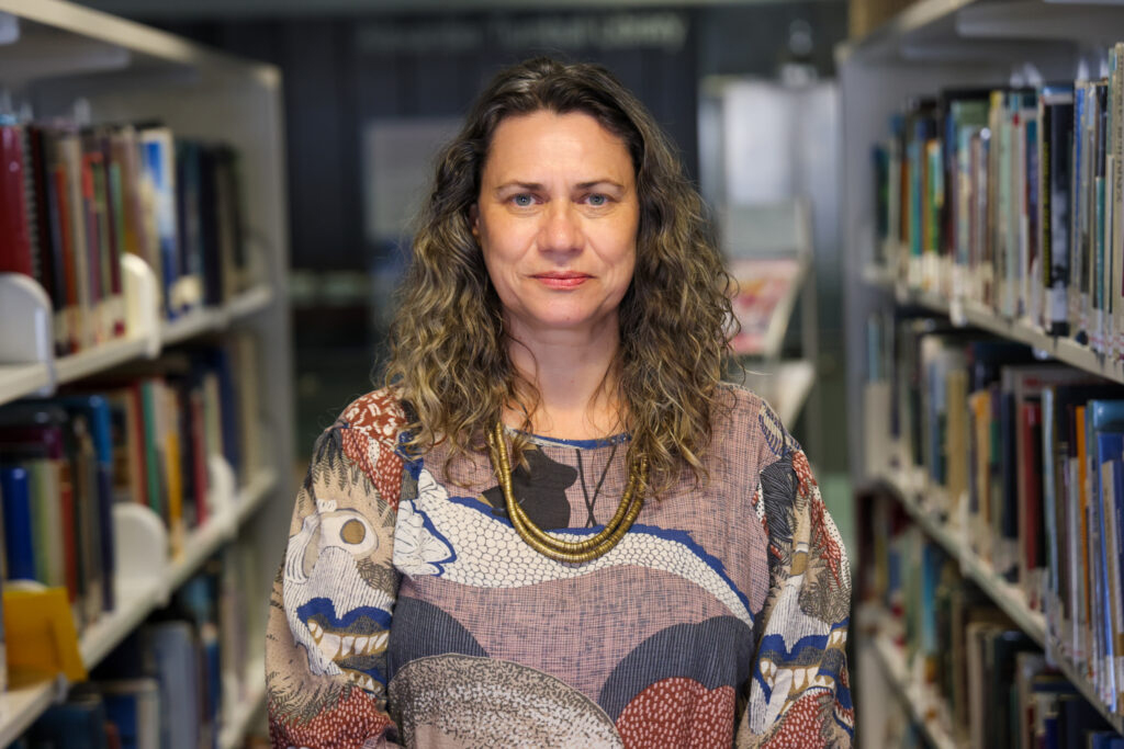 The top half of a woman with long wavy hair. She is standing in between two rows of shelves in a library.