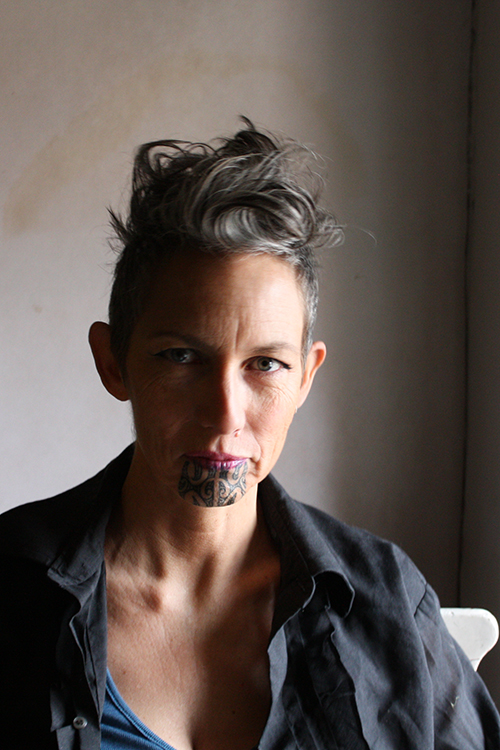 Head and shoulders of a woman with a dramatic hair cut and a moko kauae on her chin.