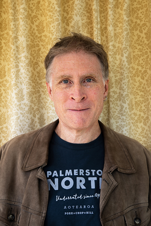 Head and shoulders of a man looking at the camera. He is standing in front of a floral yellow curtain.