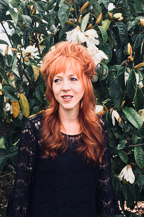 Head and shoulders of a woman with long red hair. She is standing in front of a flowering tree.