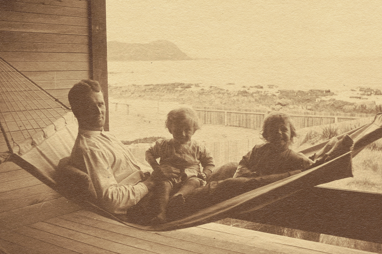 A sepia photo of a man in a hammock with two small children on his lap.