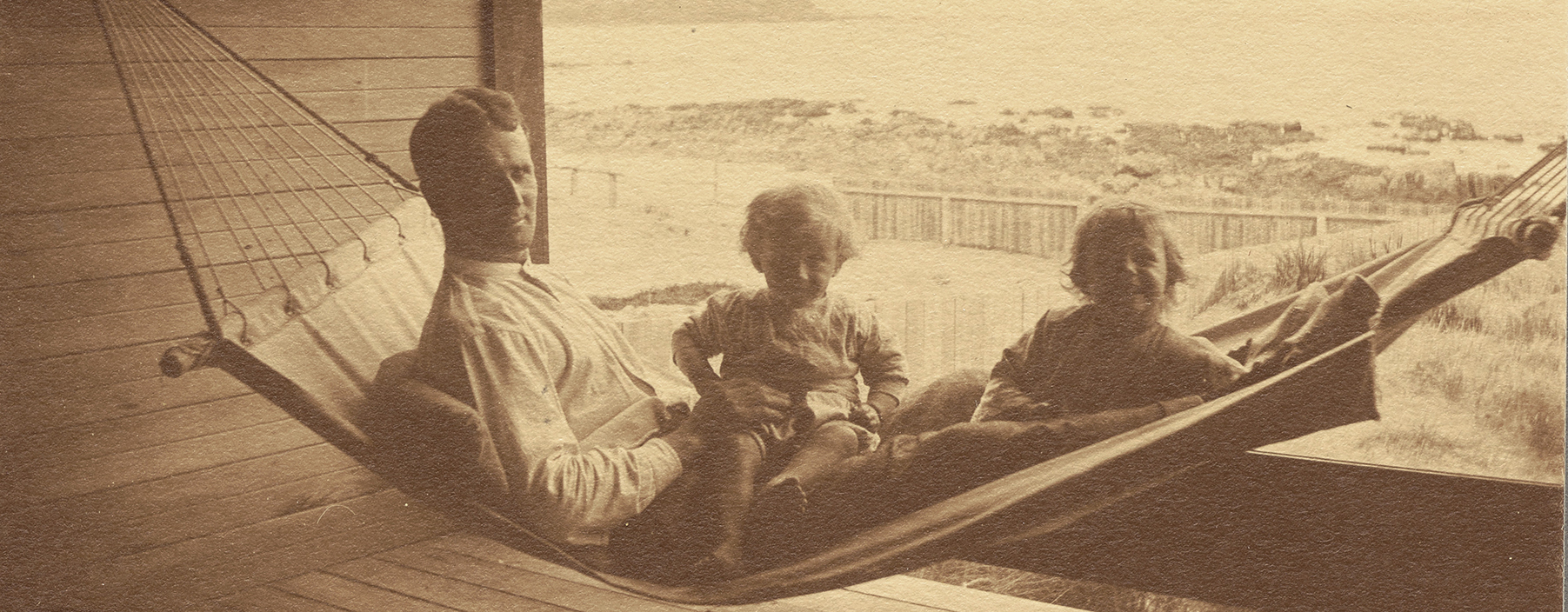 A sepia photo of a man in a hammock with two small children on his lap.