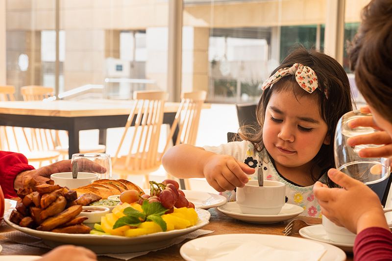 -	A child wearing a bow on their head stirs their tea. A platter of fruit, chips, and dips sits on the table in front of them.