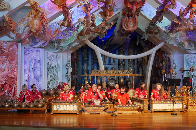 Musicians in vibrant red attire perform gamelan music on ornate bronze percussion instruments. They are seated on a stage surrounded by beautifully carved and colorful Māori patterns and sculptures on the walls and ceiling