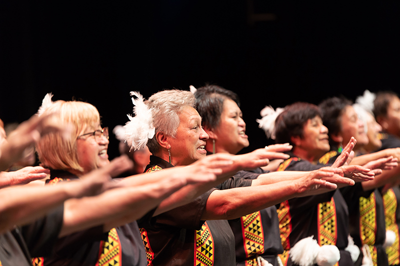 Several older women are performing on stage. They are all facing the same direction with their arms up at shoulder level.