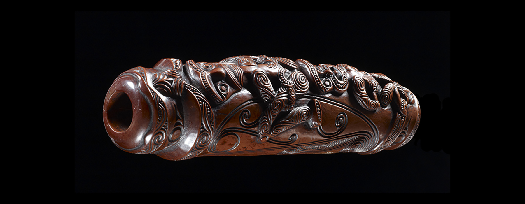 This large carved wooden koauau or flute is phallic in shape, with the ends shaped in the form of the glans penis. Either side of the stop-notes are carved in the scrolling pattern, although one side is still incomplete. The underside is carved with two humanistic ancestral figures.