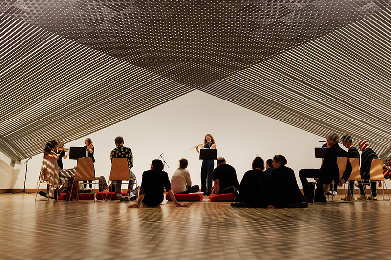 A woman is playing a flute in front of people sitting and standing under the Mataaho artwork Takapau.