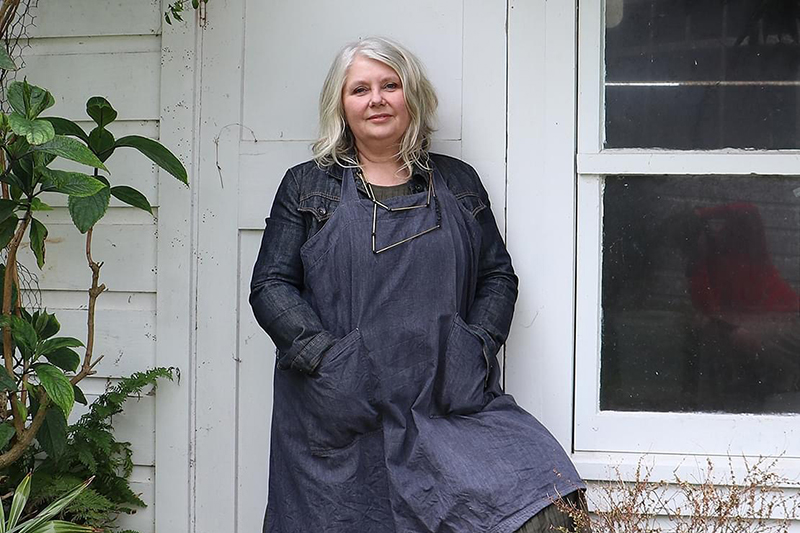 A woman with grey hair and a long denim outfit is leaning against a house.