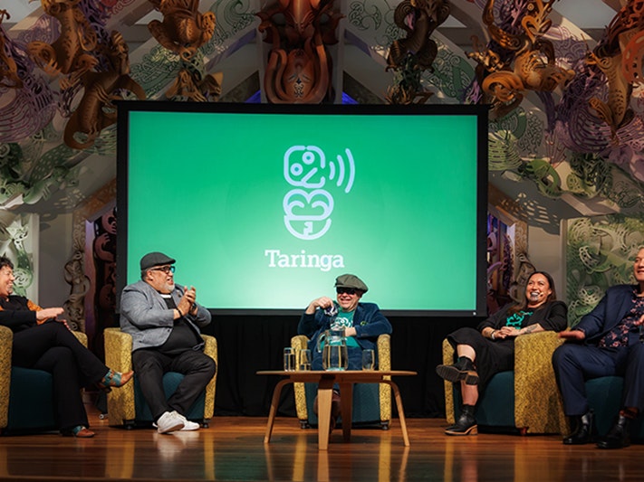 Taringa Live Podcast: Te Maori, 2024. Photo by Jo Moore. Te Papa (247569) Five people are sitting on a stage in a half circle. There is a large green screen behind them with a stylised design of a hei tiki.