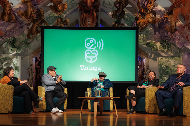 Five people are sitting on a stage in a half circle. There is a large green screen behind them with a stylised design of a hei tiki.