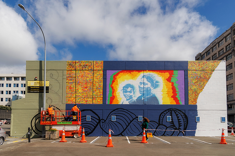 A photo of a mural being painted on a wall outside on a sunny day.