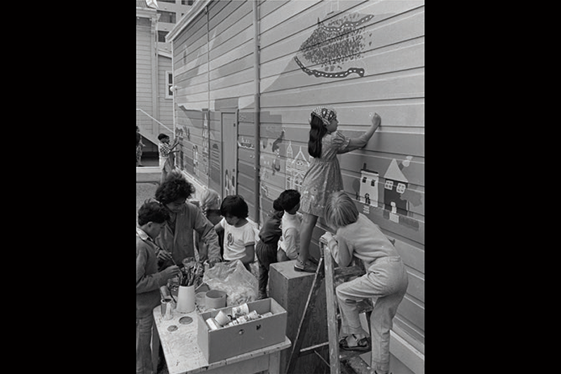 A black and white photo of children painting a mural on a wooden building