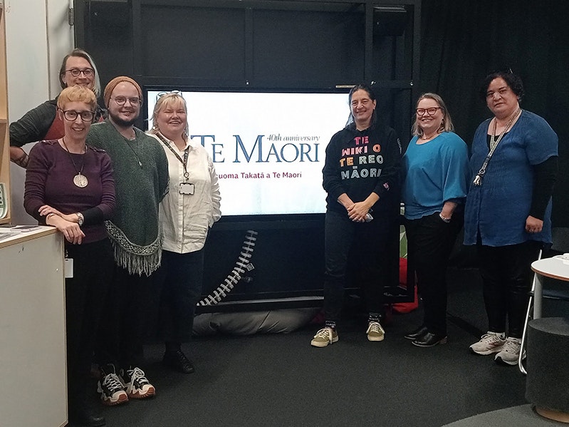 Happy Wikipedians old and new. Photo by Elise Goodge, CC BY 4.0 Seven people standing in front of a screen with the words Te Māori 40th anniversary on it.