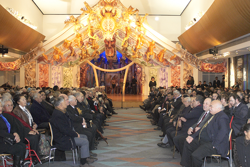 A large indoor spacec with a lot of people sitting in rows facing each other. At one end there is a carved and colourful stage.