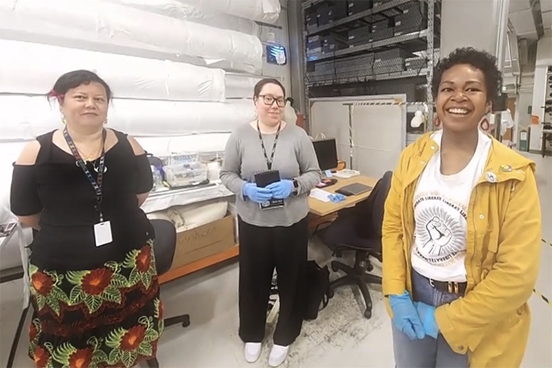 Three women are standing in a museum storage room and smiling at the camera.