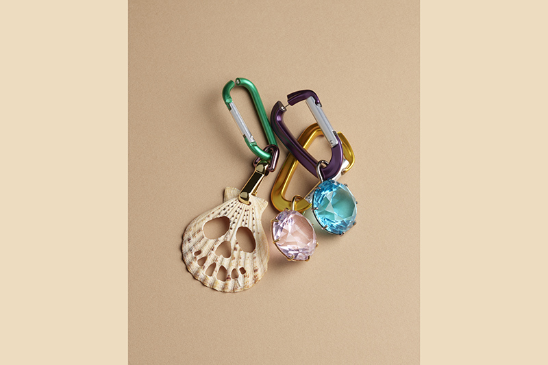 A set of colorful carabiners is attached to decorative charms, including a shell-shaped piece with skull cutouts and two large gemstone-like pendants, one pink and one blue, set against a beige background. 