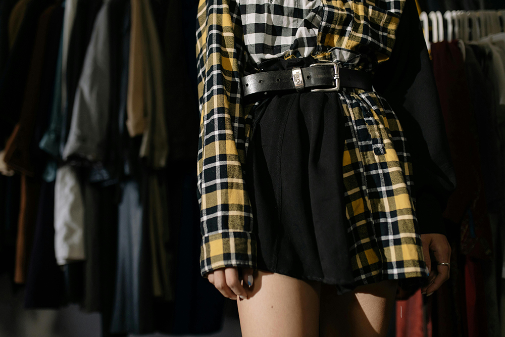 A person wearing a belted outfit with mismatched plaid patterns, featuring black and yellow checks, stands in front of a blurred clothing rack background. 