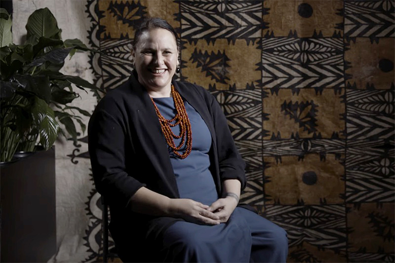 A woman is in a blue outfit and sitting in front of a large tapa cloth and smiling at the camera.