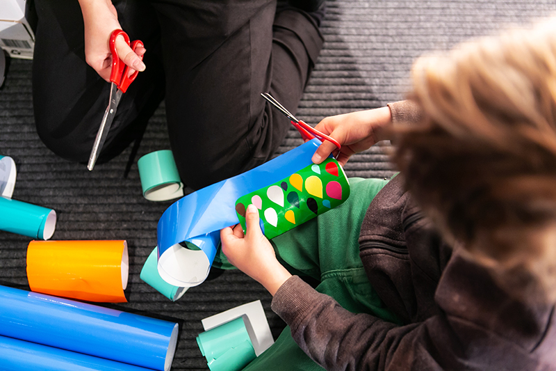 A view of the top of a head of a child cutting wrapping paper and tape with scissors.