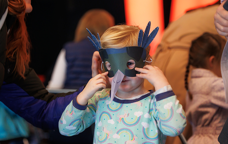 A child is holding a mask up to their face. The mask has two eyes and a beak, and paper feathers poking up from the top. There are other blurred out children in the background.