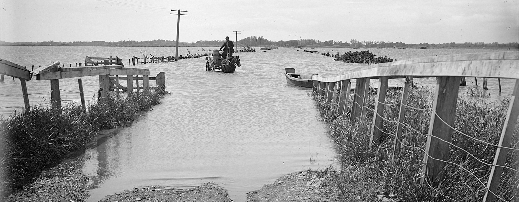 A black and white photo of a man on a horse and cart crossing a flooded roadway.