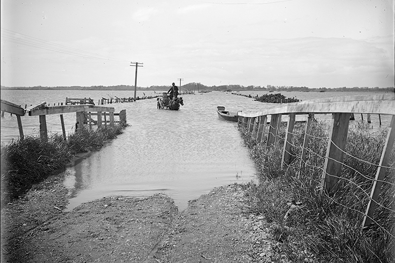 A black and white photo of a man on a horse and cart crossing a flooded roadway.