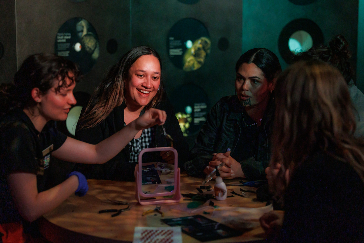 A photo of 5 adults sitting around a table in low lighting. The facilitator leading the activity demonstrates a creative action with a mirror. The 2 participants facing the camera are smiling