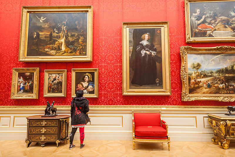 A person in black and red tights is in a gallery with large paintings on a red wall.