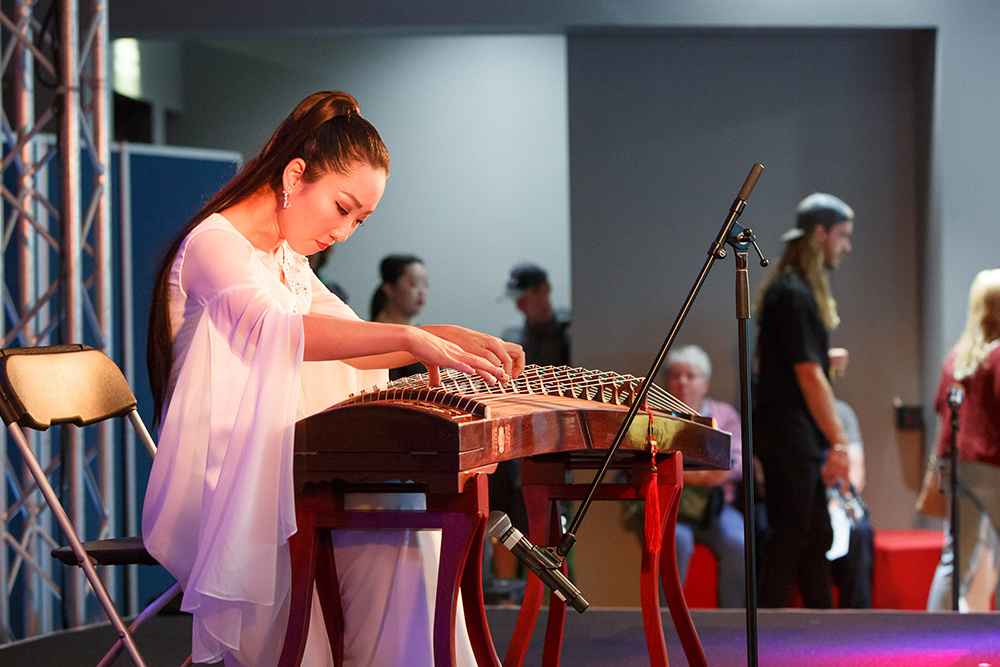 A woman is playing a stringed instrument on a stage. There are people milling in the background.