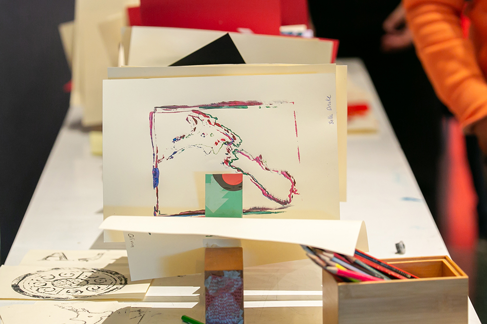 A table with some linocut prints propped up in a file. The one in the foreground has a stamp of the map of New Zealand in several different colours overlapping themselves.