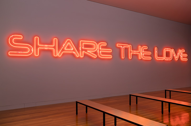 Bright red neon sign reads "SHARE THE LOVE" on a wall above wooden benches.