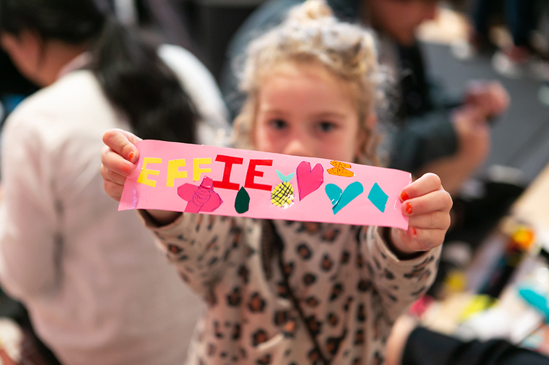 A child is holding up a strip of paper they have created. It has the word Effie on it and heart stickers and drawings.